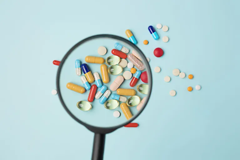 Comprimés, pilules et gélules pharmaceutiques placés sous une loupe, sur un fond bleu pastel