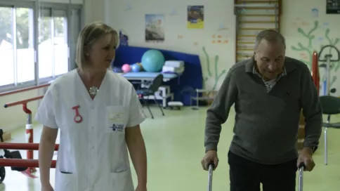 Une personne âgée en béquille est accompagnée par une soignante