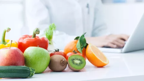 fruits et légumes posés sur une table avec en fond un médecin en blouse blanche devant son ordinateur
