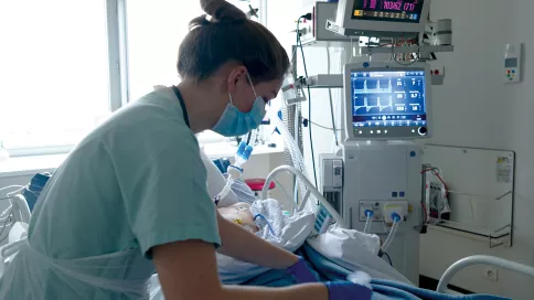 une soignante porte un masque chirurgical et des gants bleu. elle s'occupe d'une personne en réanimation. on voit en fond les moniteurs vitaux